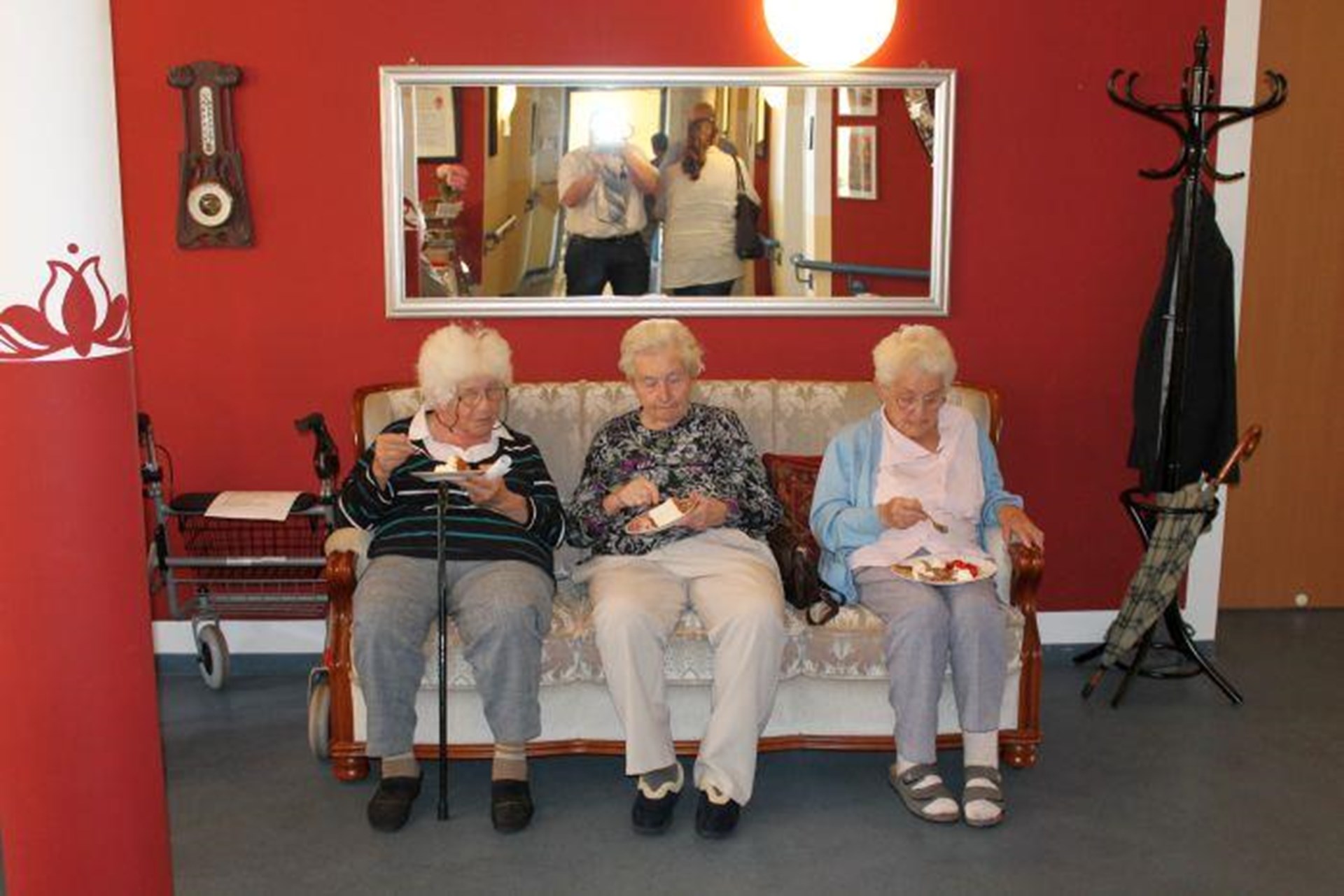 In einem rot gestrichenen Zimmer steht unter einem breiten Spiegel ein altes Sofa. Auf ihm sitzen drei Damen und essen Kuchen.